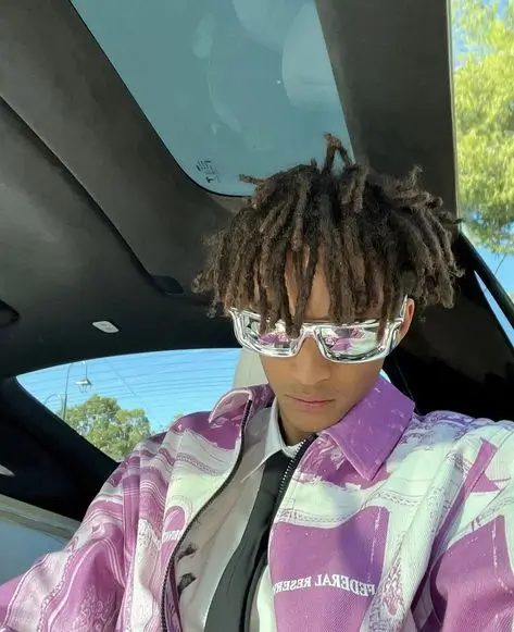 jaden wearing a white shirt with black tie and light blue denim jeans but switching up his outerwear by wearing a purple and white puffer jacket