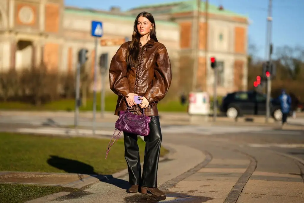 Woman standing outside wearing a leather jacket carrying a purple Balenciaga classic city bag.