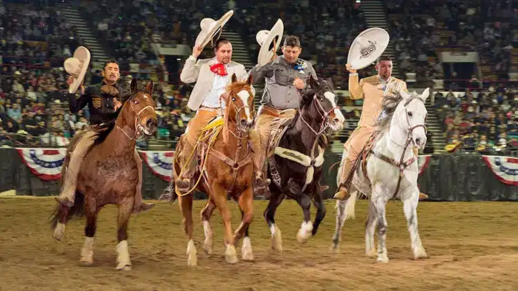 Five Mexican cowboys on horseback, dressed in traditional charro attire, performing at a rodeo. They are raising their wide-brimmed hats in a lively and vibrant display, with an audience in the background.