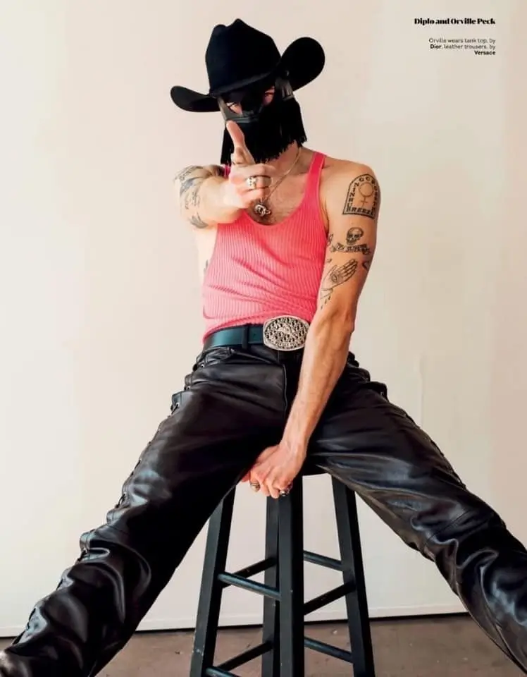 Man wearing a black cowboy hat and face covering, black pants with a statement belt and a pink tank top.