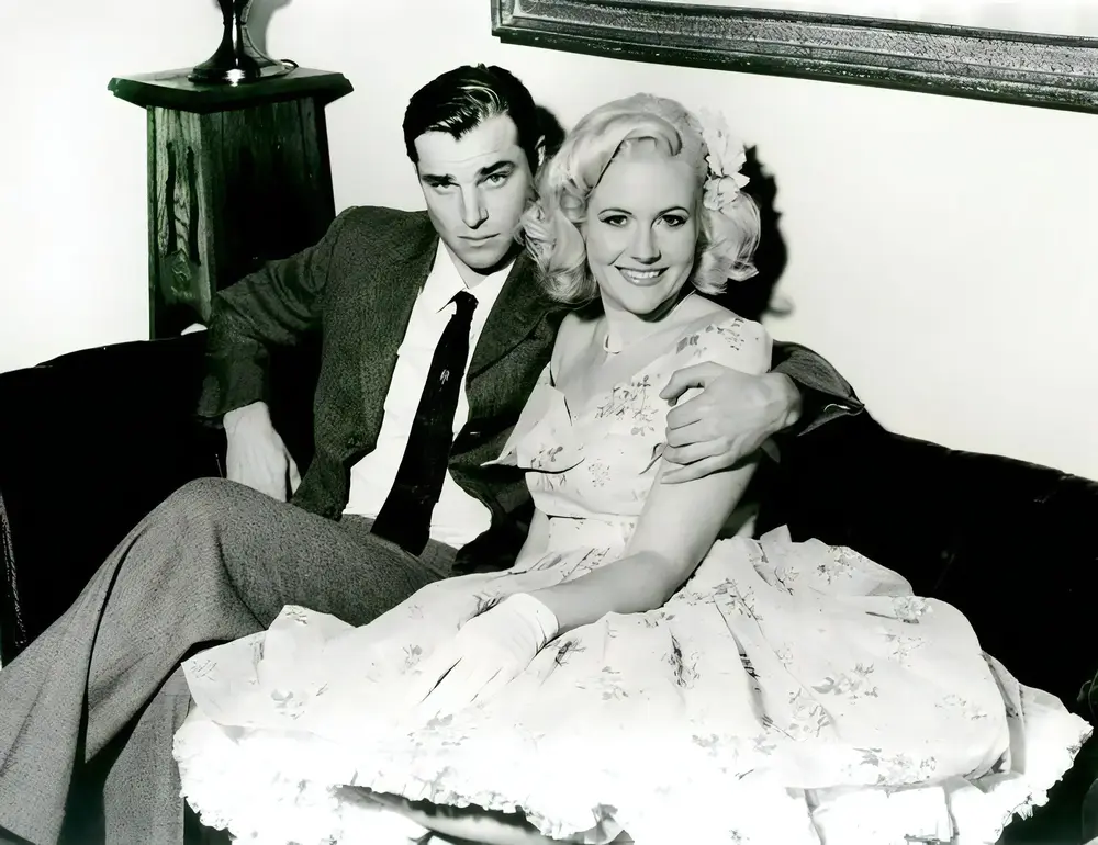 A black and white image of a young couple from the 1950s sitting on a couch. The man is dressed in a suit and tie, and the woman is wearing a floral dress with gloves and a flower in her hair. They are smiling and looking at the camera.