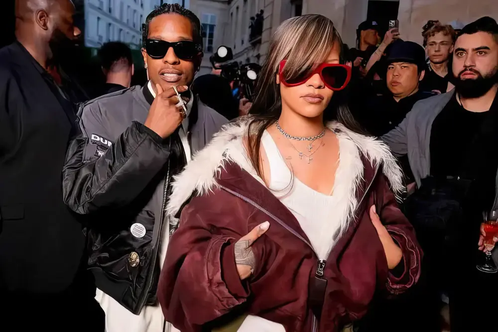 Rihanna and A$AP Rocky attending Paris Fashion Week. Rihanna is wearing a burgundy jacket with white fur trim and large red sunglasses, while A$AP Rocky sports a black bomber jacket and sunglasses. The background features a bustling crowd and Parisian street scenery.