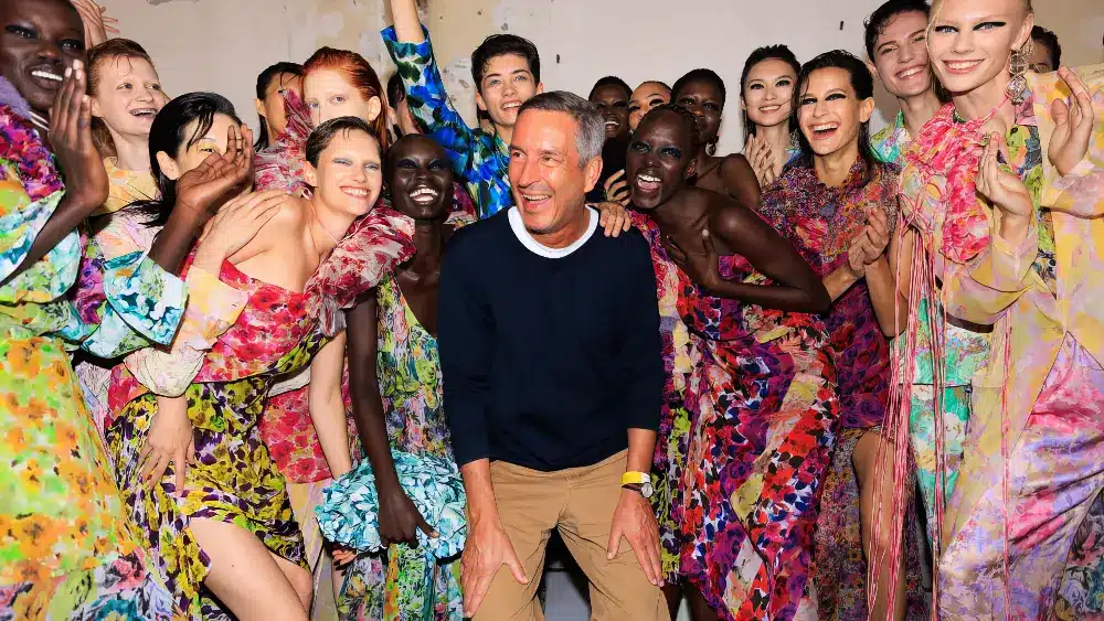 Dries Van Noten laughing and surrounded by models dressed in vibrant, floral outfits from the SS23 collection.