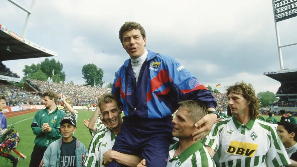 Otto Rehhagel held aloft by Werder Bremen players in his distinctive Reebok tracksuit, celebrating a victory with cheering fans in the background.