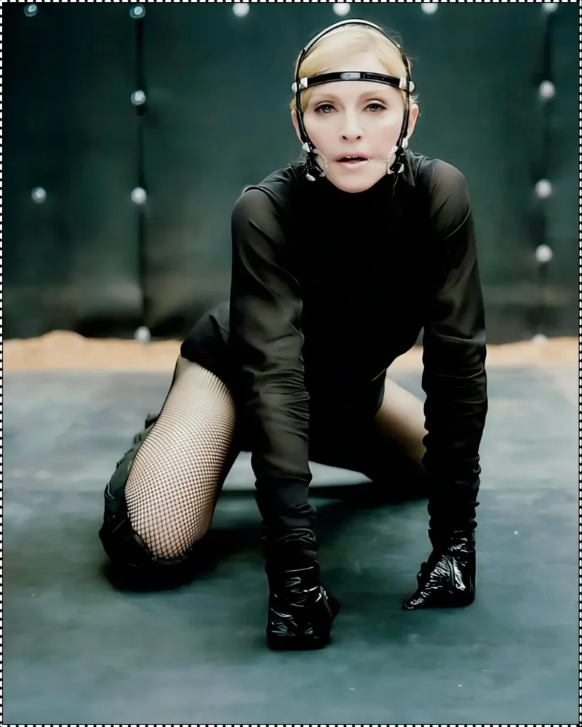 A powerful fashion image by Steven Klein of Madonna dressed in a black bodysuit, fishnet stockings, and gloves, kneeling on the ground with futuristic headgear, exuding strength and intensity.