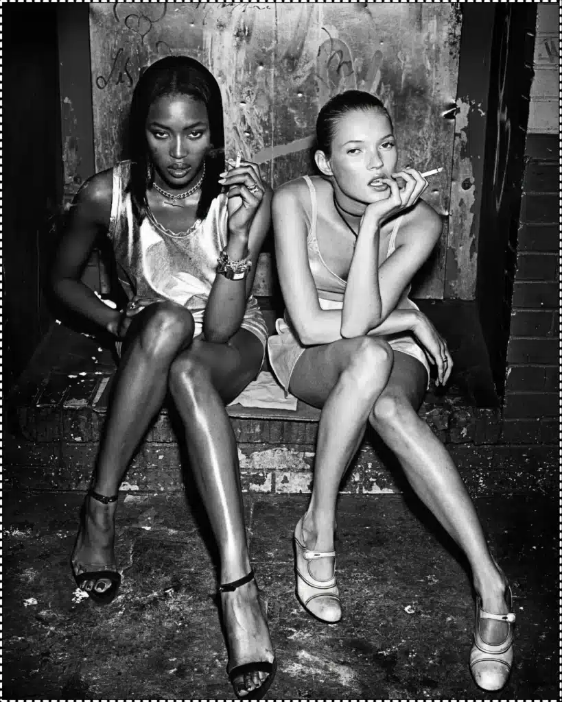 Black and white Steven Klein captured image of Naomi Campbell and Kate Moss sitting beside each other with cigarettes in their hands staring seductively at the camera