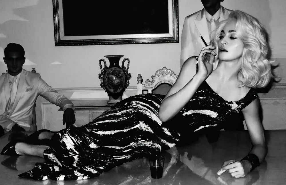 Black and white image of Madonna laying down on a hotel room table holding a cigarette with two hotel staff in suits behind her. The image was take by Steven Klein